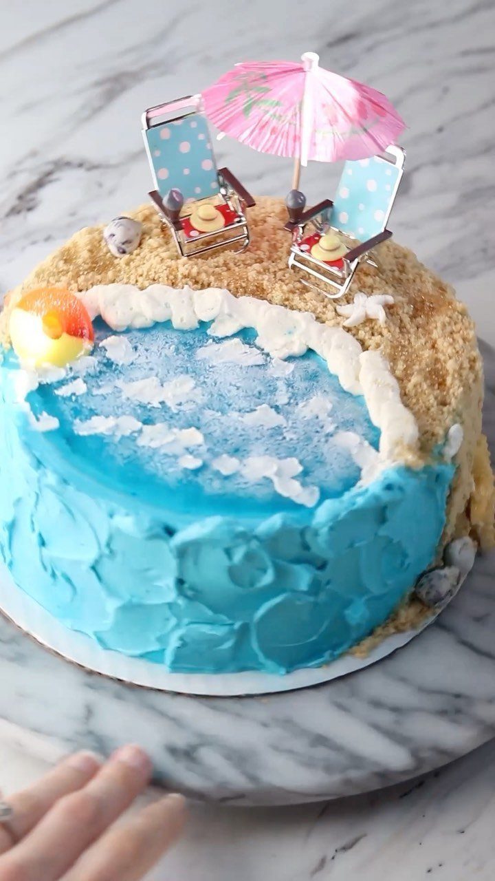 26 Beach Cake Ideas with Blue Ocean Frosting to Sailboats 20 Beach themed cake with blue frosting, sandy crumbs, and a pink drink umbrella.