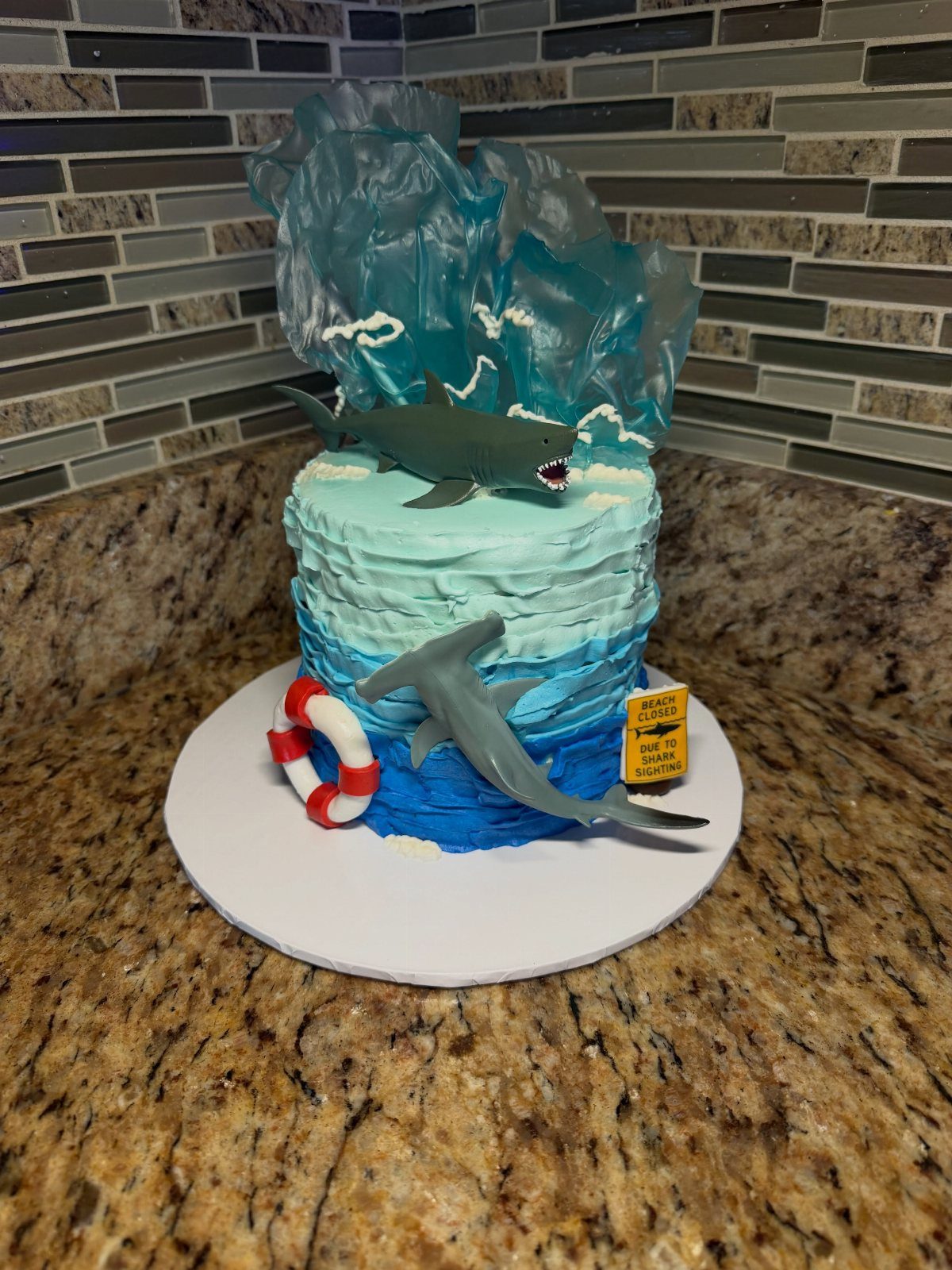 26 Beach Cake Ideas with Blue Ocean Frosting to Sailboats 21 Beach cake with blue frosting, a gelatin wave, and two grey gummy sharks.