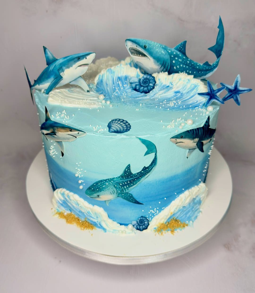 26 Beach Cake Ideas with Blue Ocean Frosting to Sailboats 10 Blue ombre cake decorated with sculpted sharks, white frosting waves, and blue seashells.