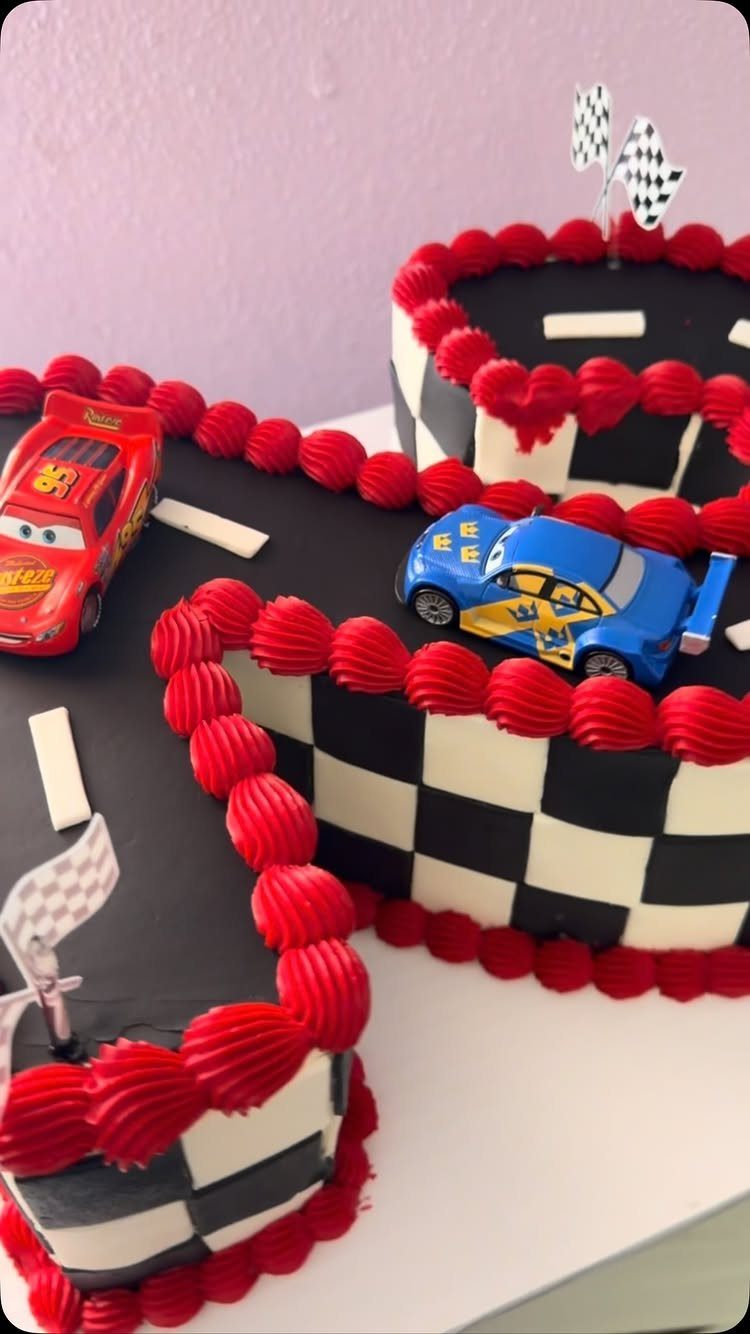 Race track shaped cake with red borders, checkered patterns, and toy racing cars.