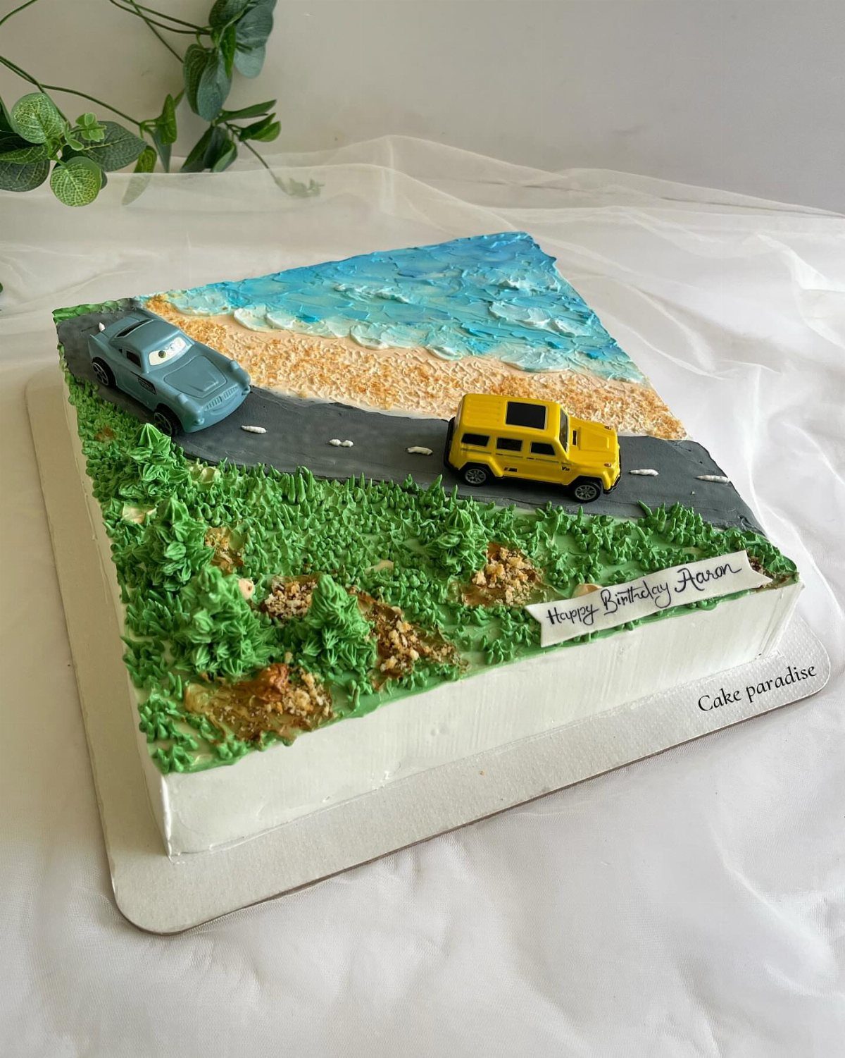 Square cake decorated as a coastal road with small toy cars and beach scenery.