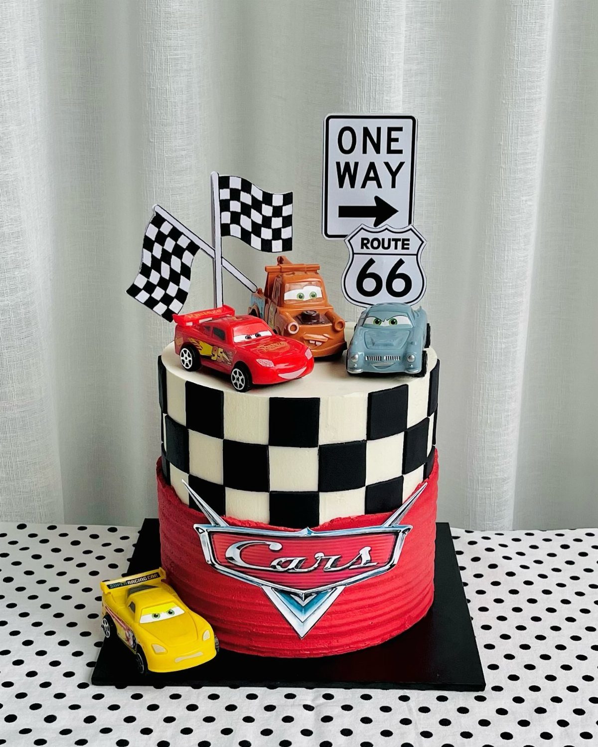 Two-tier car themed birthday cake with checkered patterns and toy racing cars.