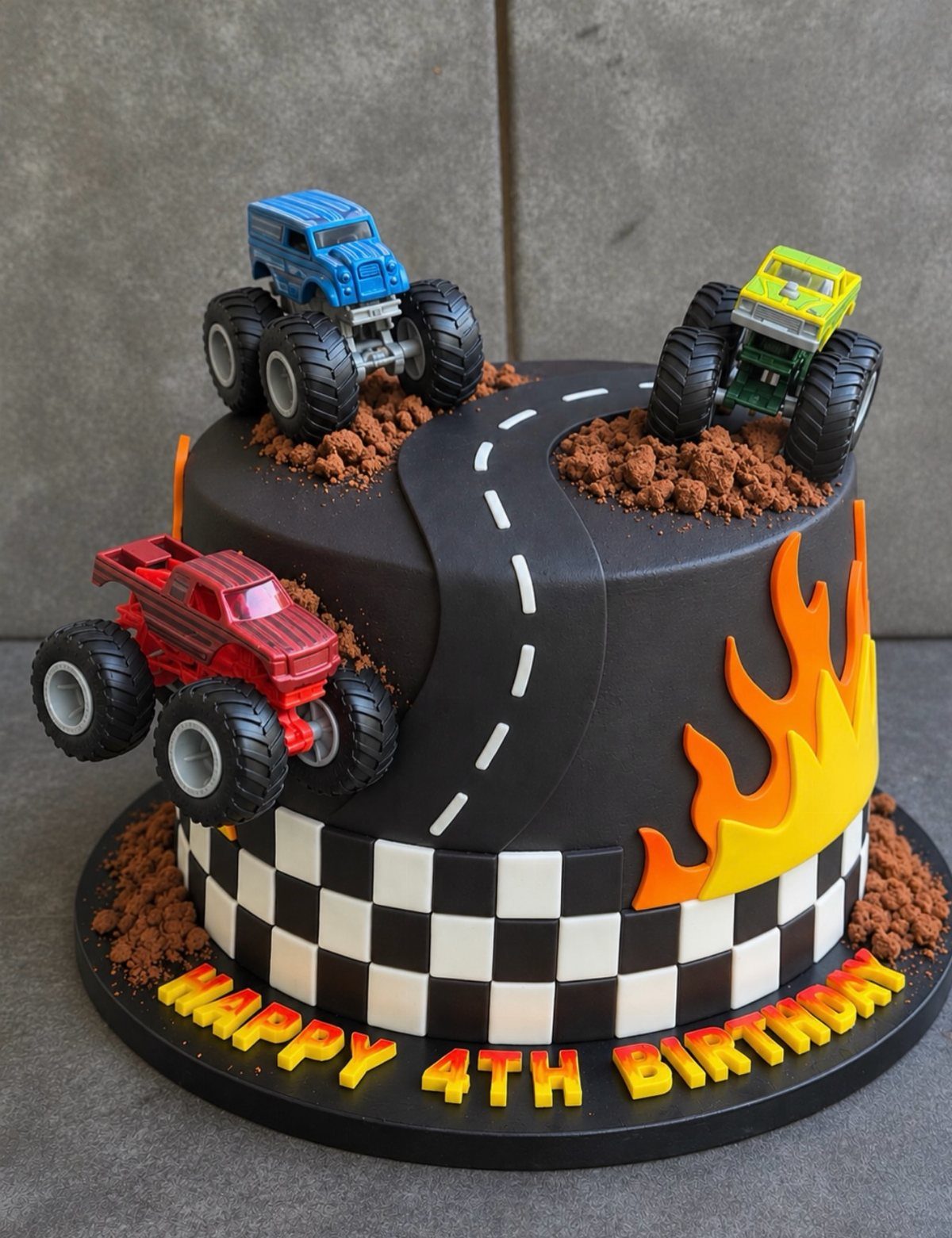 Two-tier black monster truck cake with toy trucks, dirt, and checkered racing flags.