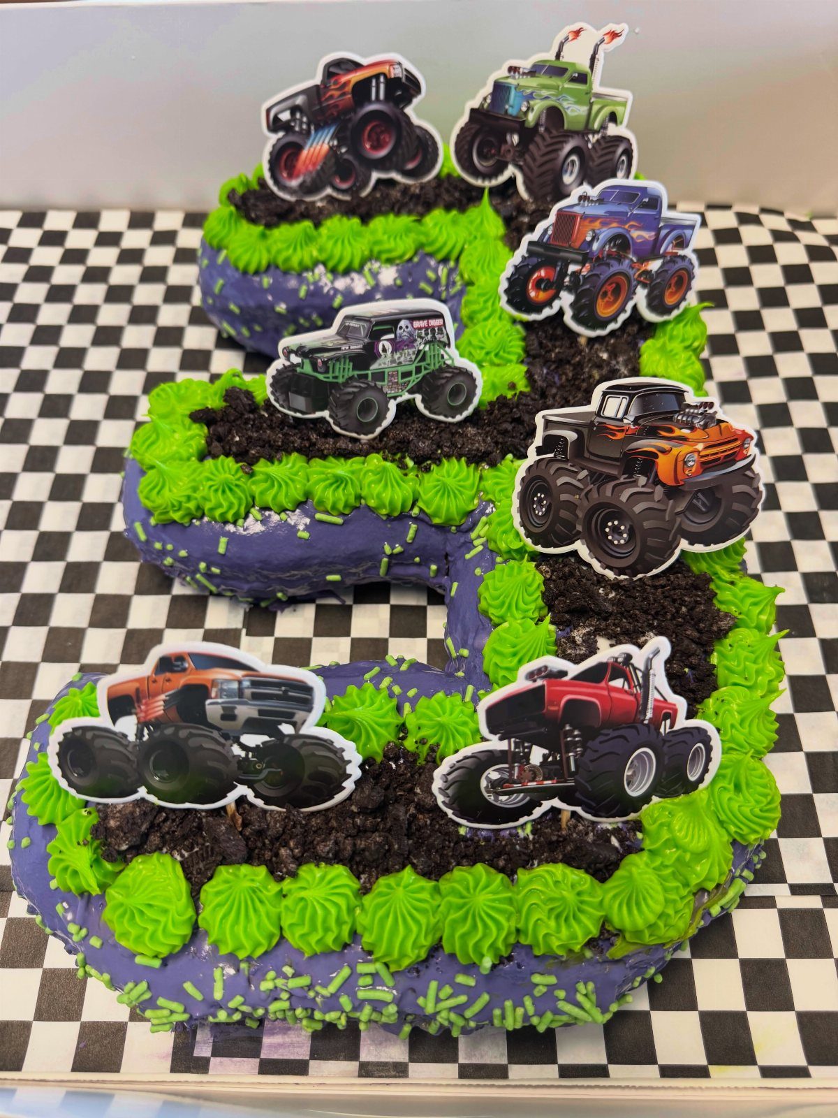 Number 3 shaped cake with purple frosting, green swirls, and monster truck toppers.