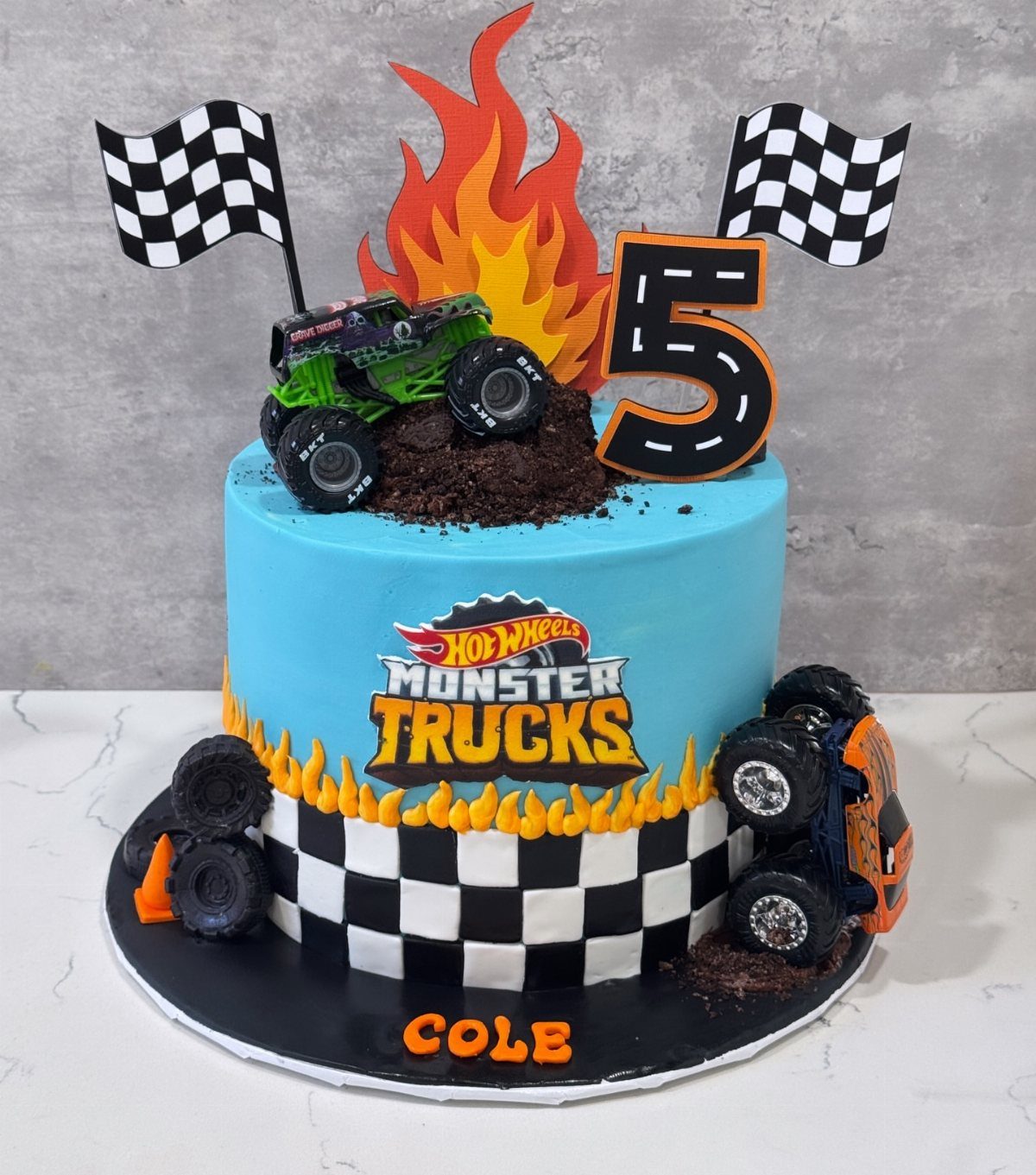Monster truck themed birthday cake with blue frosting, checkered patterns, and toy trucks.