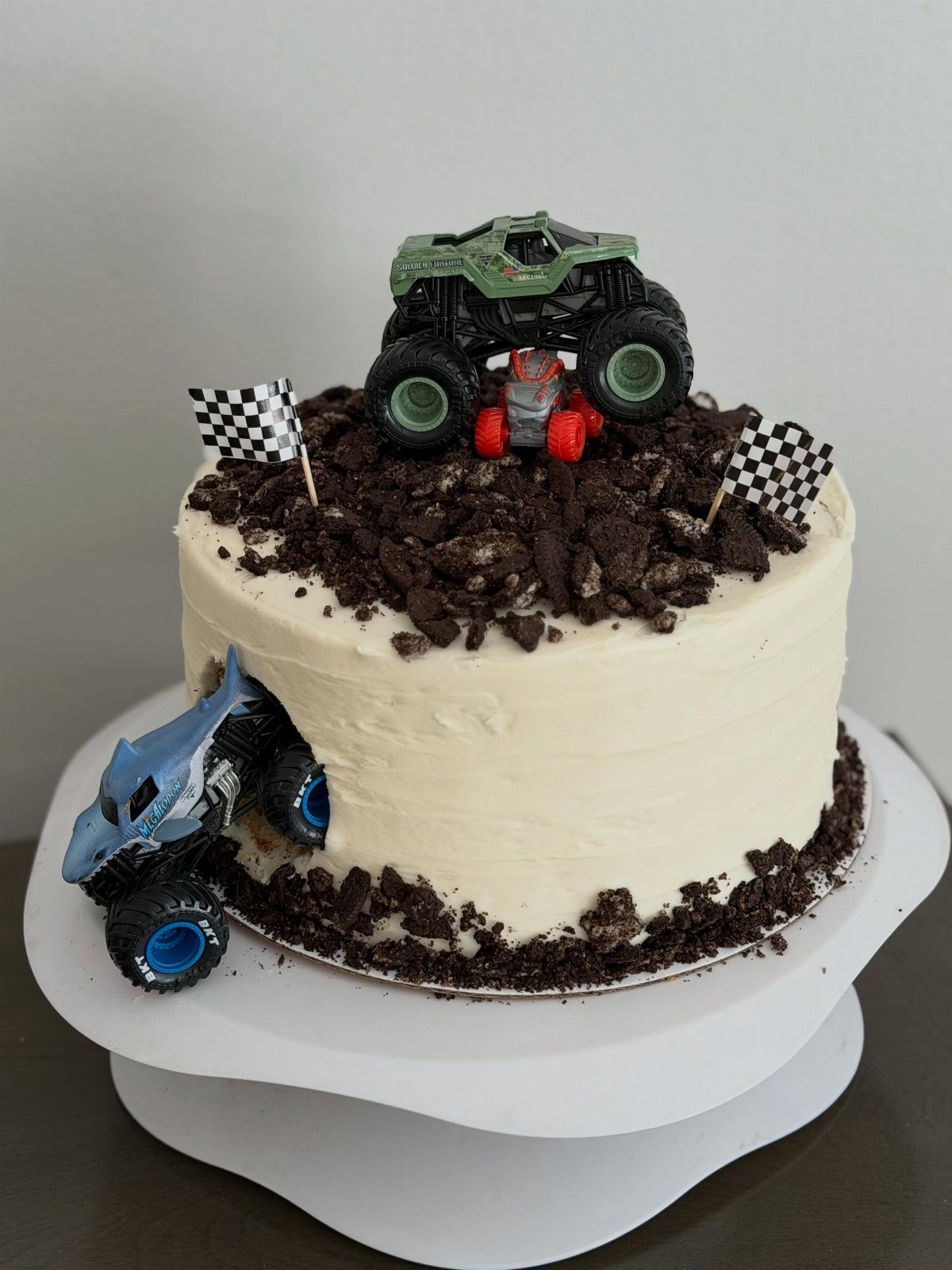Monster truck cake with white frosting, chocolate dirt, and toy trucks and flags.