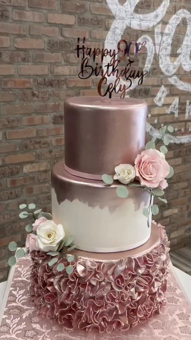 Three-tier cake with rose gold ruffles, metallic finish, and pink floral decorations.