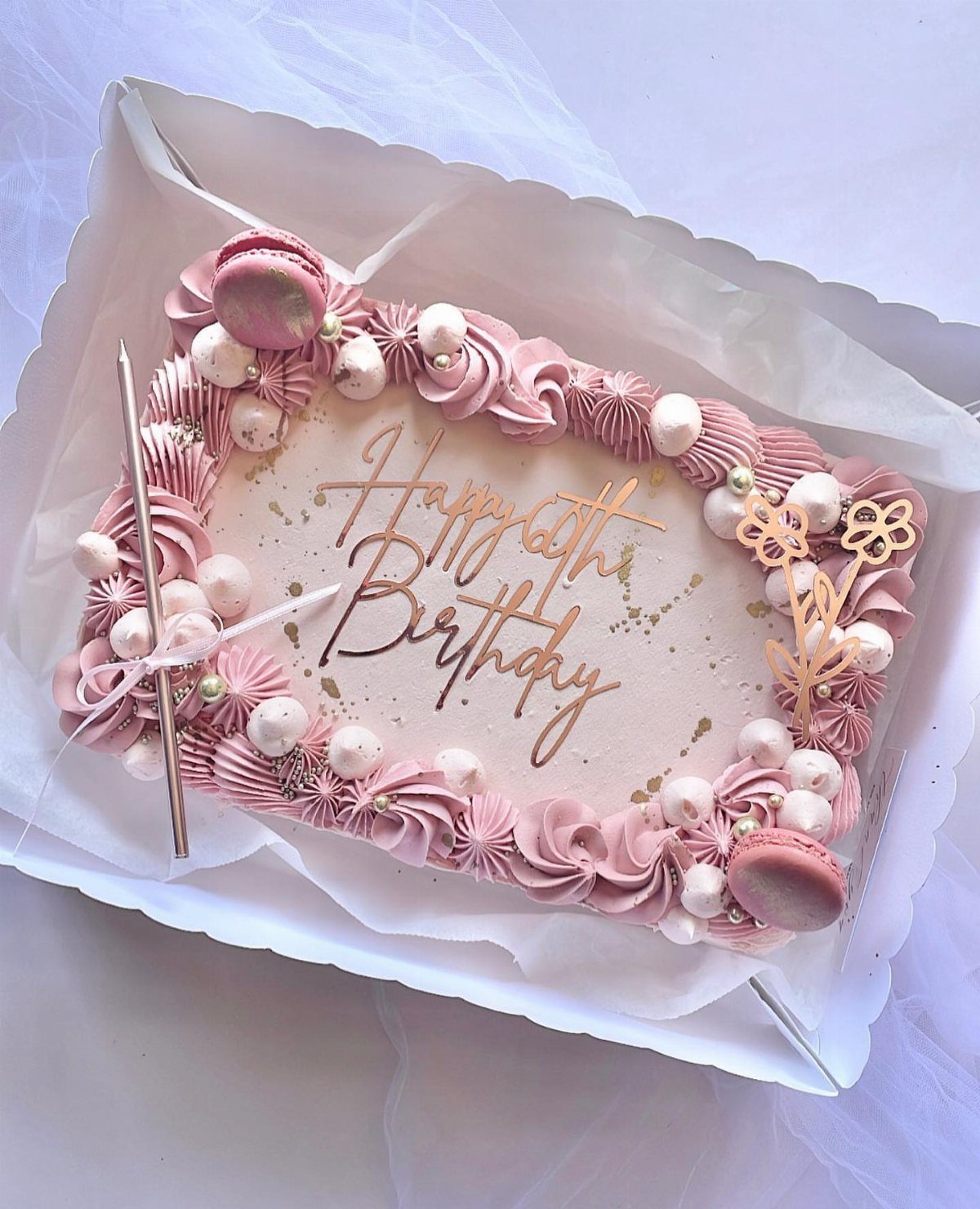 Pink rectangular birthday cake with rose gold lettering, macarons, and frosting swirls.