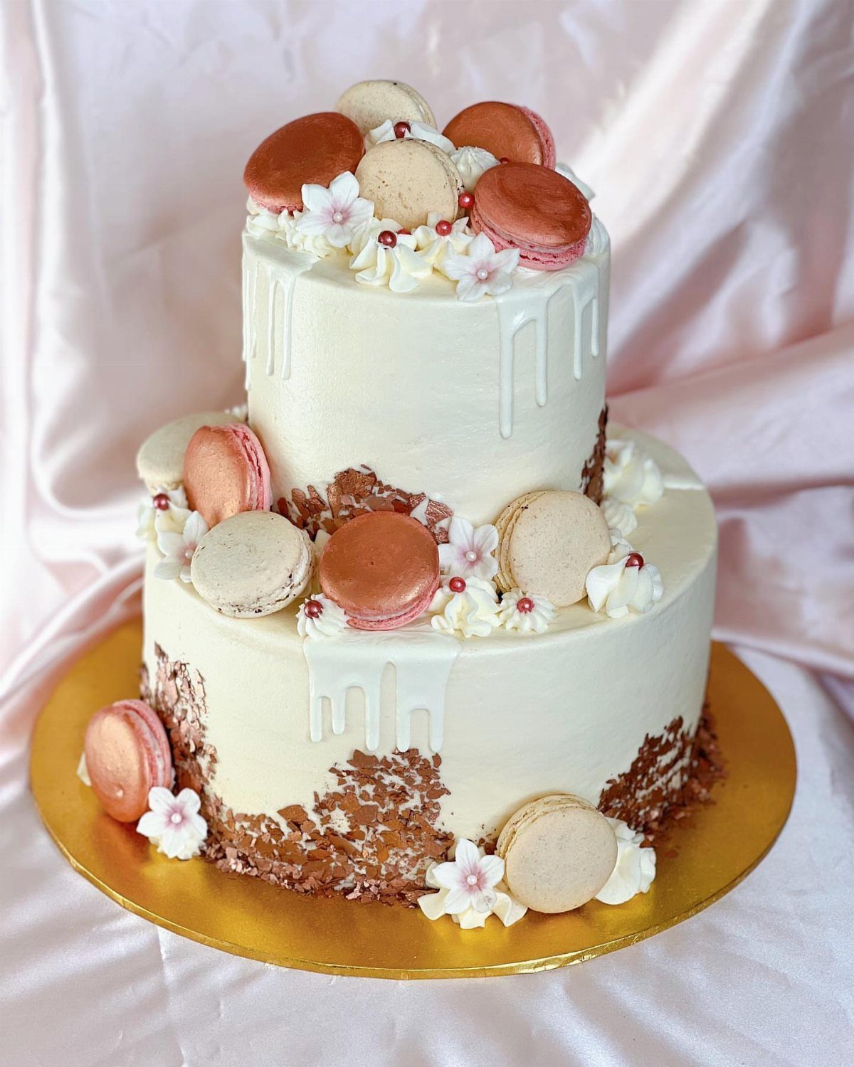 Two-tier white drip cake decorated with rose gold macarons and small white flowers.