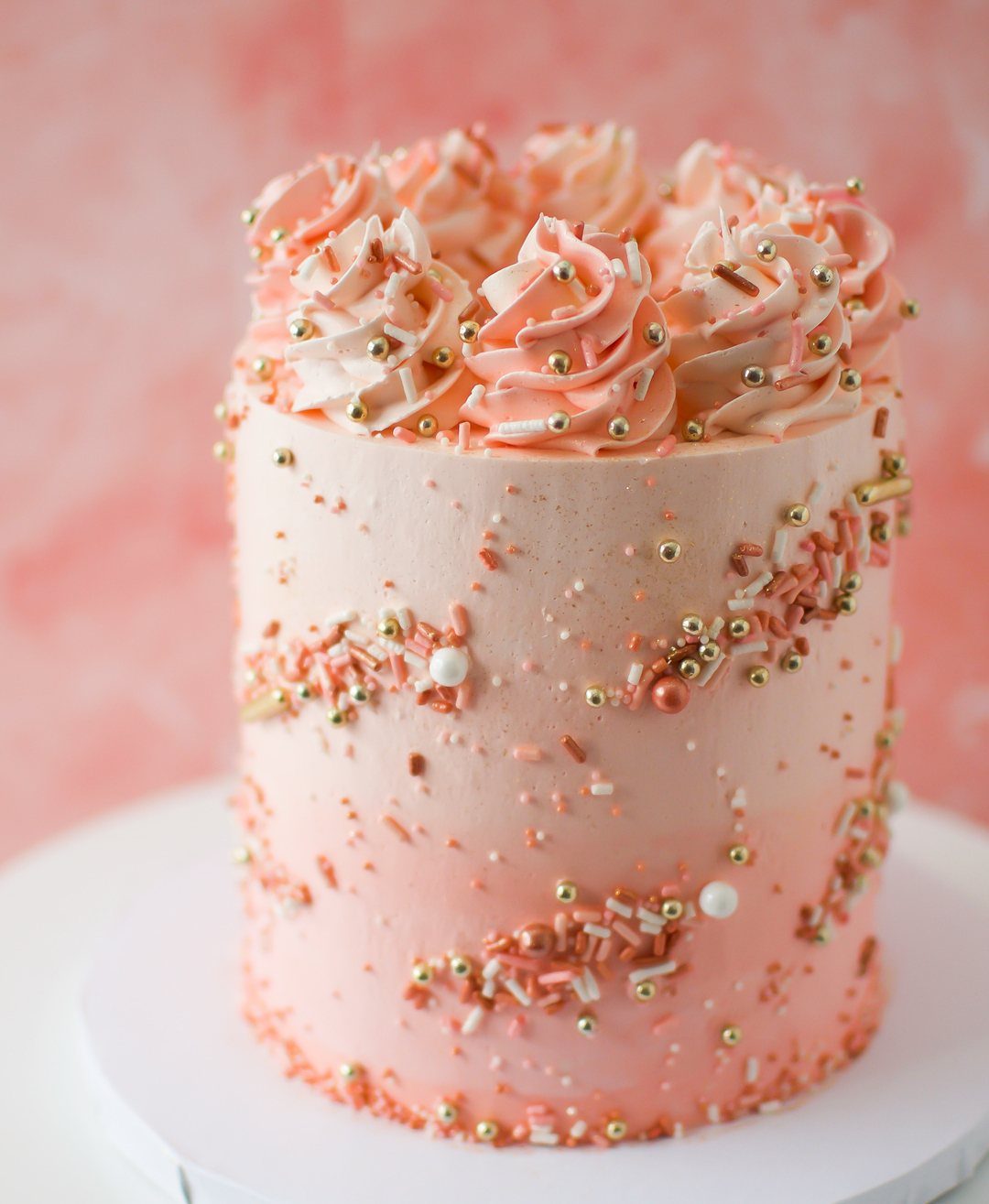 Peach cake with rose gold sprinkles and metallic pearls on a white stand.